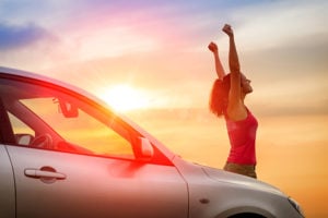 Female driver beside car raising arms facing towards the sunset